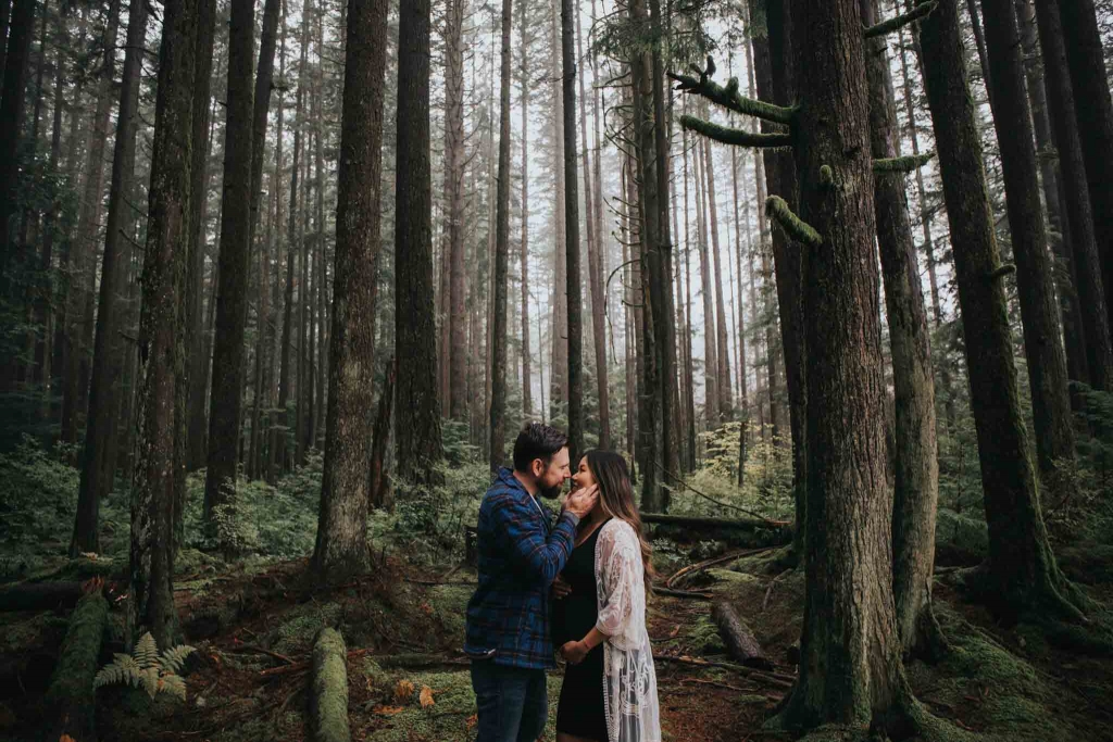 pregnant couple standing in forest kissing